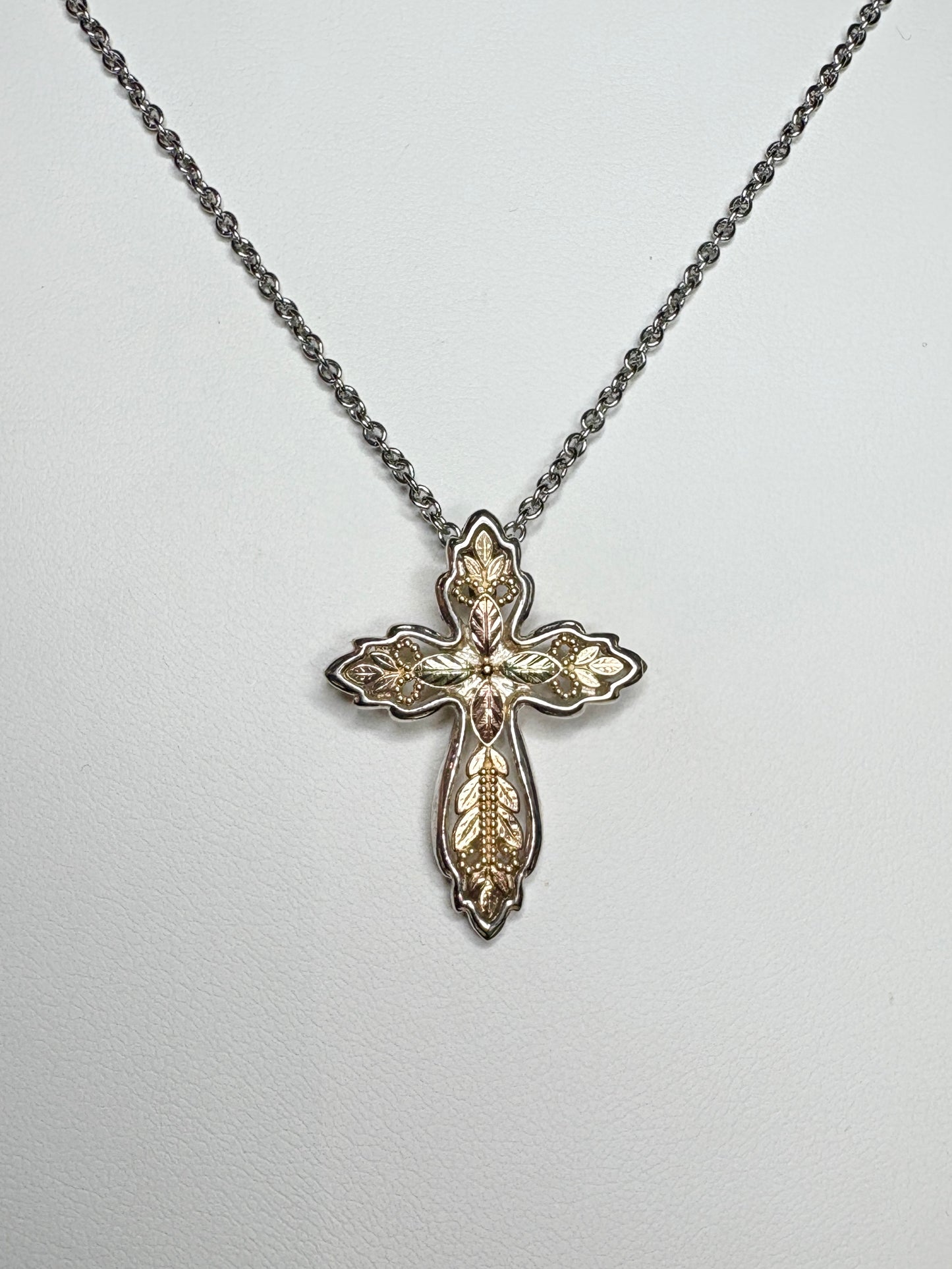 Black Hills Gold ornate cross pendant with green and rose gold leaves on a yellow gold base with a sterling silver frame.