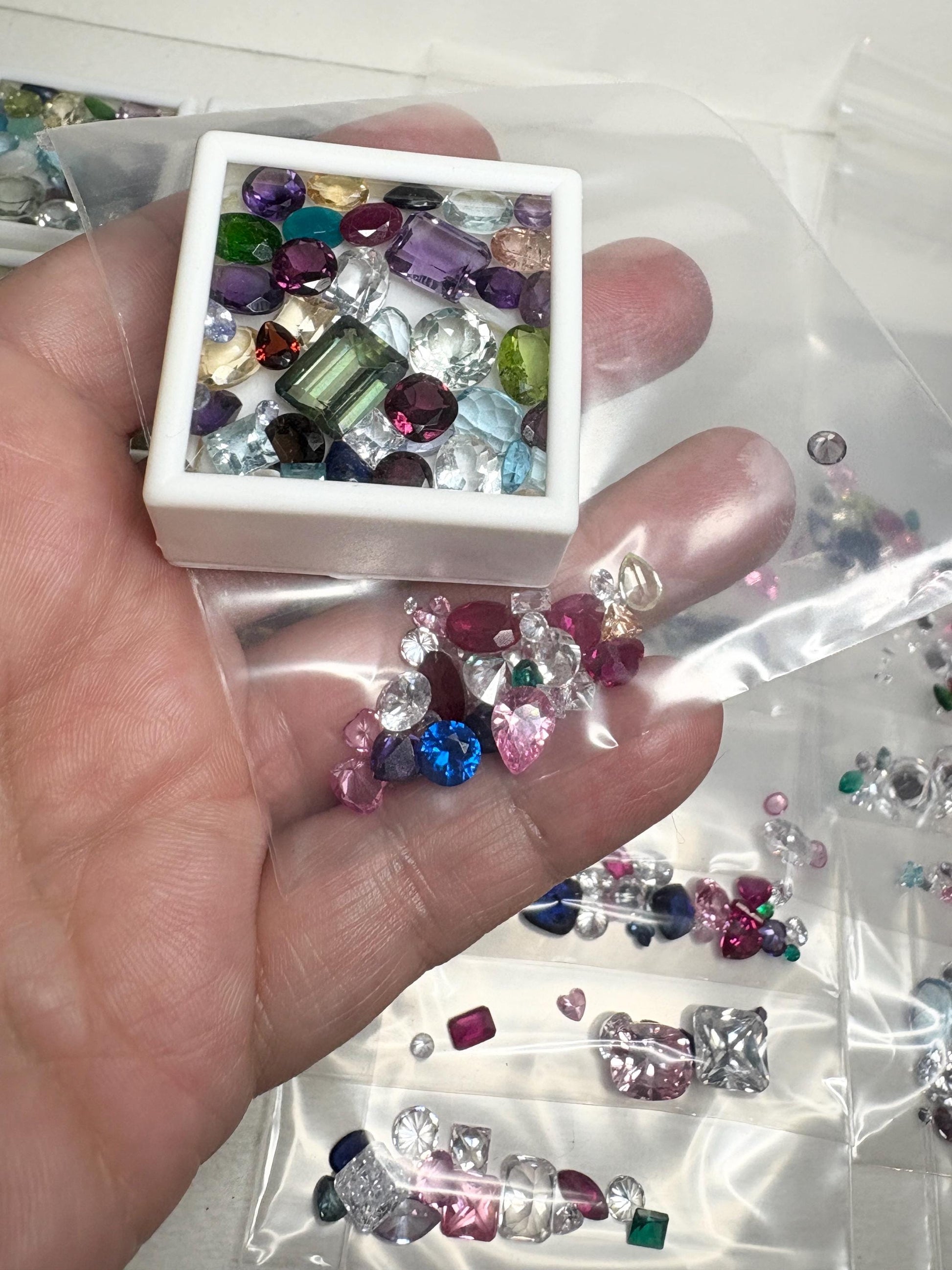 A hand holds a white tray of mixed faceted natural gemstones and a plastic bag of loose gemstones.